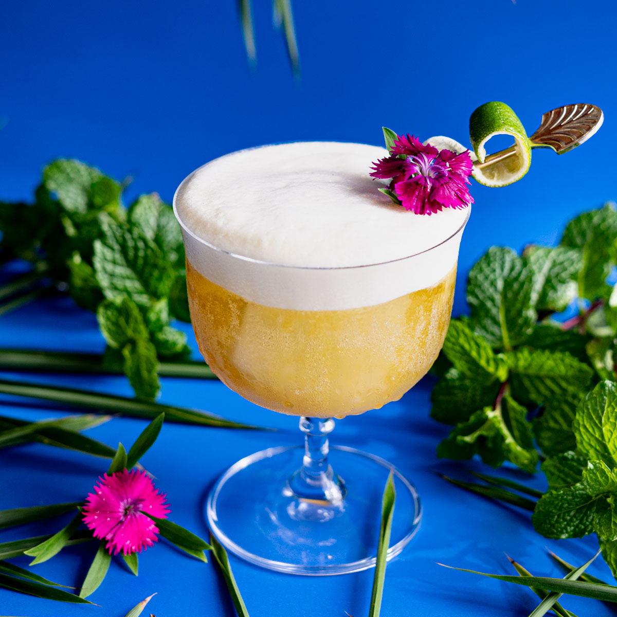 A tropical pineapple cocktail is pictured sitting on a blue background surrounded by mint leaves, bamboo leaves, and a tropical flower. The drink is garnished with a lime peel and a tropical flower.