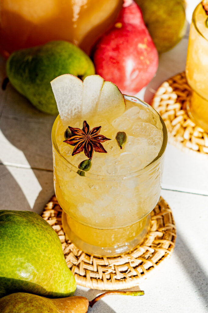 A photograph shows a Fall Rum Punch made using pears and a blend of three rums. The drink is garnished with a whole allspice pod and cardamom pods, and three slices of pears. The photo is taken by Central Florida Food and Beverage Photographer, Lindsey Neumayer.